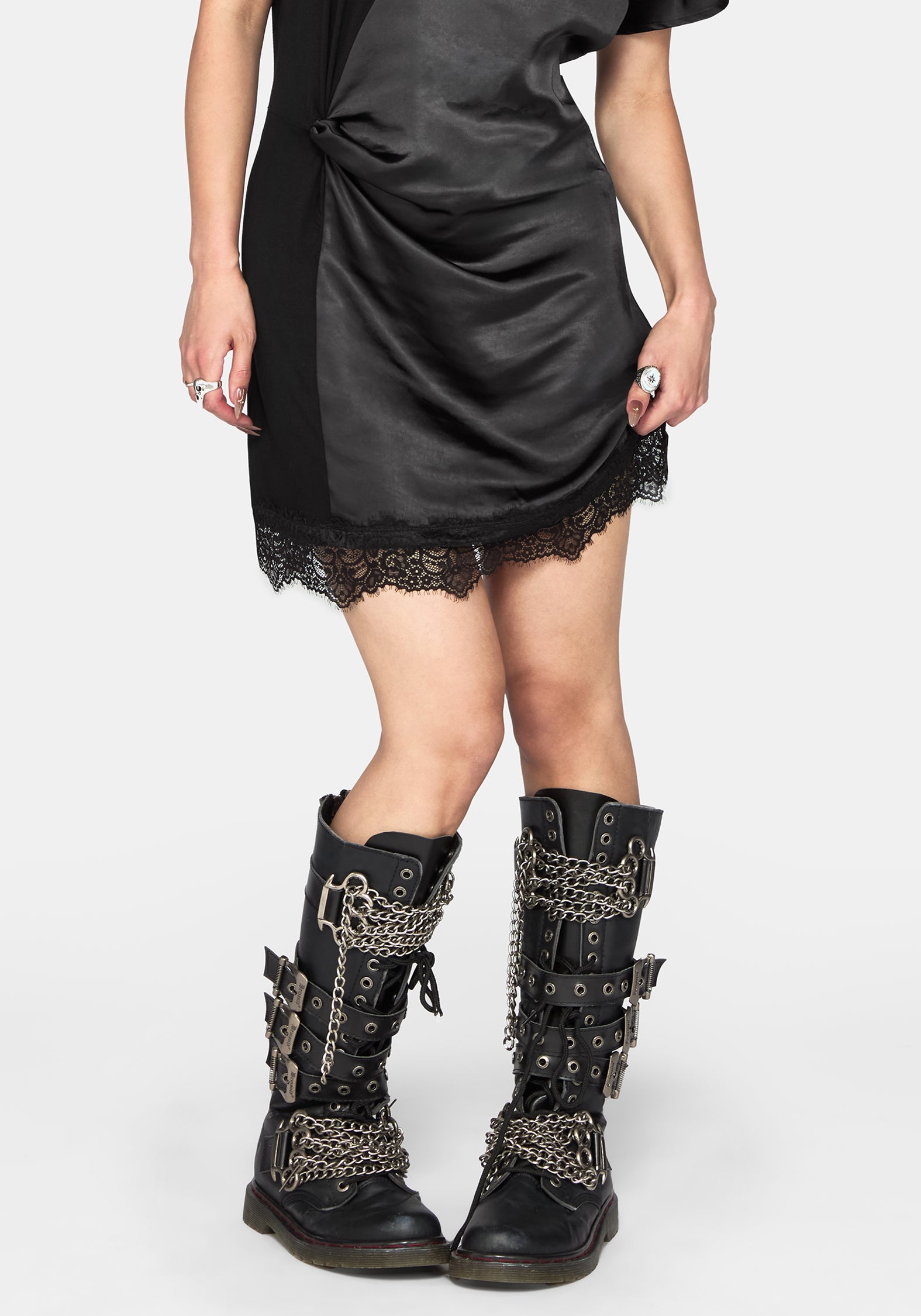 A model wearing a black satin-like mini dress with lace trim and black lace-up combat boots decorated with silver chains and buckles, photographed against a light gray studio background.