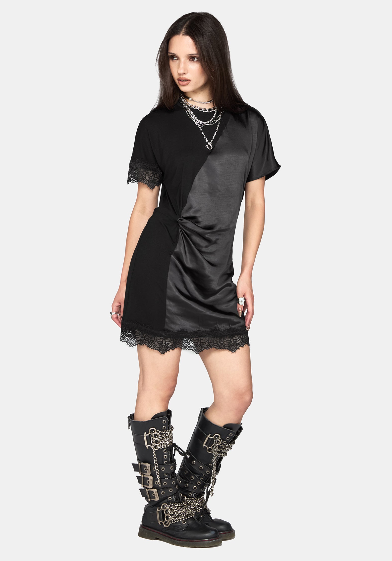 A model wearing a black short-sleeve dress with a satin-like sheen, lace trim, and a twisted detail at the waist, paired with layered silver necklaces and black lace-up combat boots, against a light gray background.