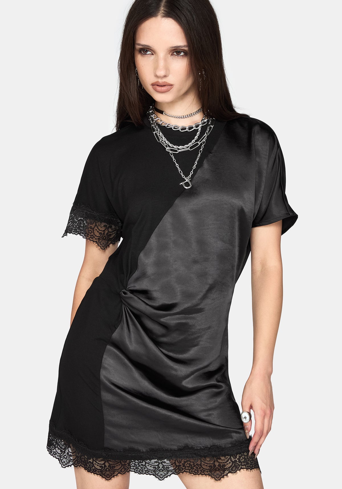 A woman modeling a black satin short-sleeve dress with lace trim on the sleeves and hem, styled with layered silver necklaces against a light gray background.