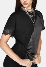 A close-up of a model wearing a black short-sleeve dress with a diagonal satin panel, lace trim on the sleeve, and a twisted detail at the waist, styled with layered silver chain necklaces.